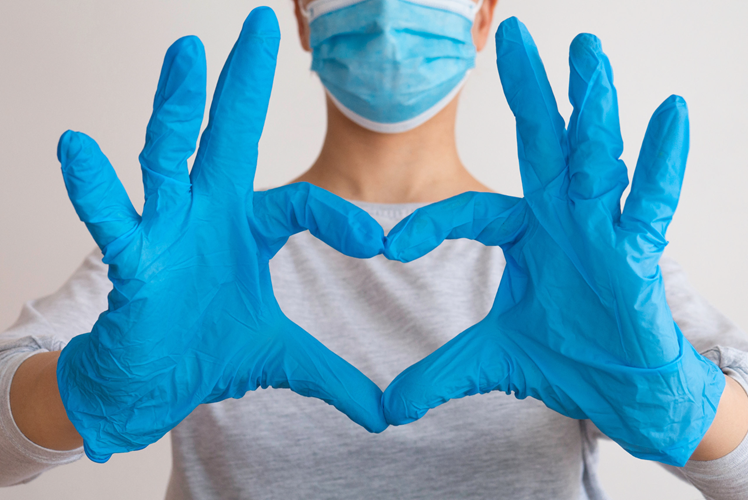 A dentist in surgical mask, wearing a light grey long sleeved shirt, wearing blud surgical gloves with their hands heald out in front of them in the shape of a love heart