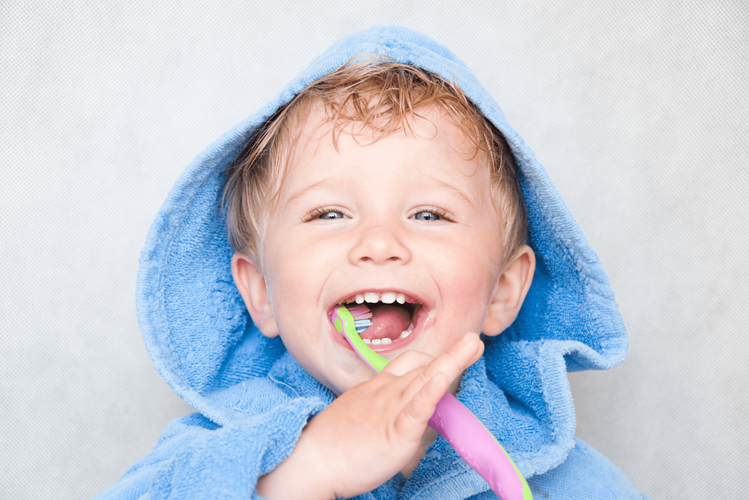 A happy smiling toddler with a blue toweling robe hood on his head, his mouth open wide holding a toothbrush in his right hand, while the toothbrush is in his mouth